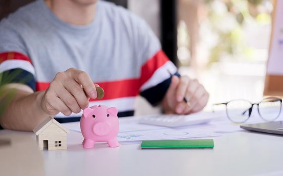 Close up young man putting coin in piggy bank and calculate savings money plan to buy property, house. Personal financial concept for own a house.