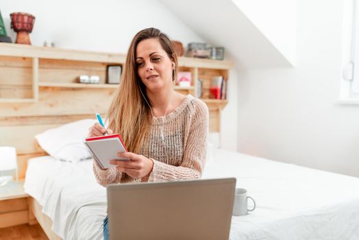 Casual Lady Taking Notes While On Call From Her Room Using A Computer With Headset, Business Woman Taking Notes Through Vedio Conference Meeting From A Laptop In Bedroom.
