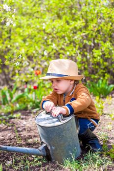 Cute little toddler boy in a hat and rubber boots is watering plants with a watering can in the garden. A charming little kid helping his parents grow vegetables. Active holidays with children