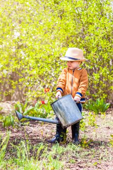 Cute little toddler boy in a hat and rubber boots is watering plants with a watering can in the garden. A charming little kid helping his parents grow vegetables. Active holidays with children