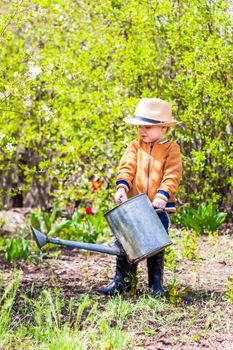 Cute little toddler boy in a hat and rubber boots is watering plants with a watering can in the garden. A charming little kid helping his parents grow vegetables. Active holidays with children