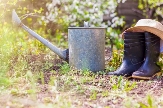 An old iron watering can, a hat, and rubber boots stand in the garden. Gardening and gardening, Outdoor activities with children 