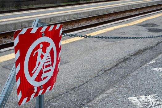 A red metal sign posted at the edge of railway tracks at a train station warns people to stay away from the track due to the danger.