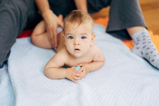Tiny baby in diapers lies with his tummy on a blue blanket holding a toy in his hands. Mom's hands hold the baby by the back. High quality photo