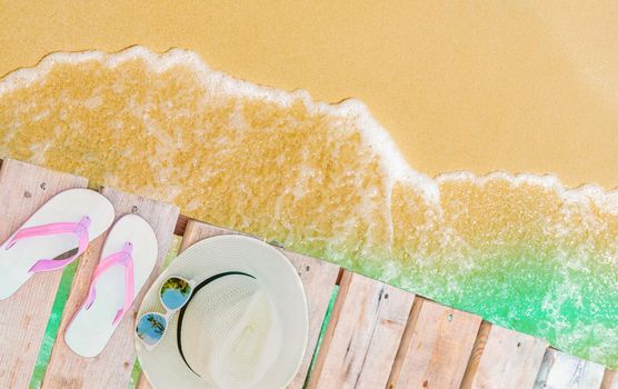 Top view of pink-white sandals, straw hat and sunglasses on wooden bridge over emerald-green sea water and golden sand beach. Summer vacation travel. Summer vibes. Coconut tree shadow in sunglasses.