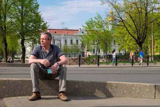 An adult caucasian senior man in a gray T-shirt sits and rests on the curb and holds a smartphone in his hand on a sunny day in the city in summer. Selective focus.