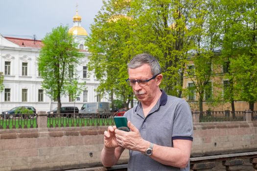 An adult caucasian senior man in a T-shirt is typing a message on a smartphone in the city in spring or summer on the embankment against the backdrop of buildings and green trees. Selective focus.