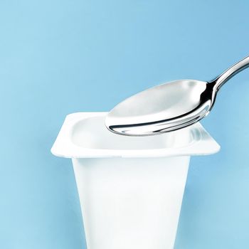 Yogurt cup and silver spoon on blue background, white plastic container, fresh dairy product for healthy diet and nutrition.