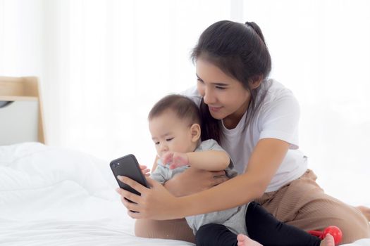 Young asian mother and little baby girl or newborn selfie with smart phone on bed in bedroom, happiness mom and daughter using phone video call at home, two people, family and communication concept.