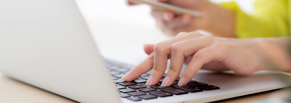 Closeup hand of young asian businesswoman working on laptop computer on desk at home office, freelance woman looking message on smart phone and typing on notebook, business and communication concept.