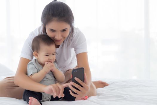 Young asian mother and little baby girl or newborn selfie with smart phone on bed in bedroom, happiness mom and daughter using phone video call at home, two people, family and communication concept.