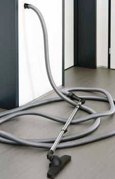 Long central vacuum cleaner hose laid on the grey floor in the room. Hose of central vacuum cleaner plugged into a wall inlet valve socket.