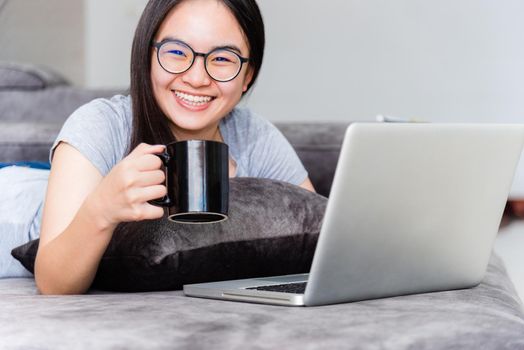 Portrait beautiful Asian young woman holding coffee cup smile and looking during a work, Cute girl teens happy on the sofa working remotely with a laptop and internet computer communication from home