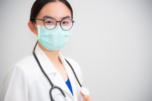 Studio portrait beautiful Asian young woman doctor with stethoscope in white uniform wear glasses and green mask to protect Corona Virus for health looking at camera on gray copy space background