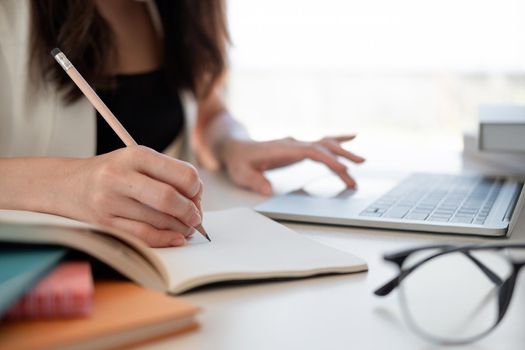 Close up woman's hands with laptop computer, notebook and pen taking notes in business office.