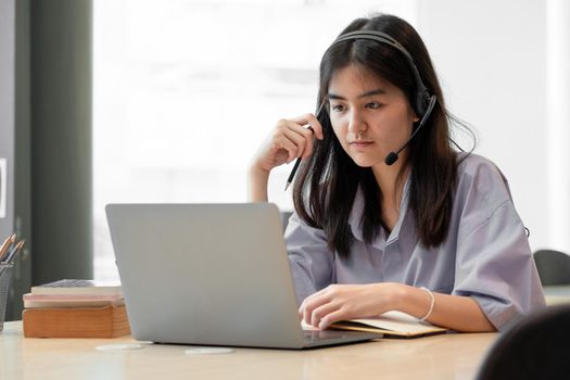 concentrated young asian girl or student looking at laptop holding book learning, serious woman working or studying with computer doing research or preparing for exam online