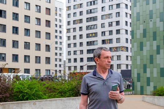 An adult caucasian man in a T-shirt stands against the background of residential buildings in the city in spring or summer and holds a smartphone in his hand. Selective focus, front view.