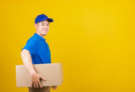 Portrait excited attractive delivery happy man logistic standing smile wearing blue t-shirt and cap uniform holding parcel box looking to camera, studio shot isolated on yellow background, side view