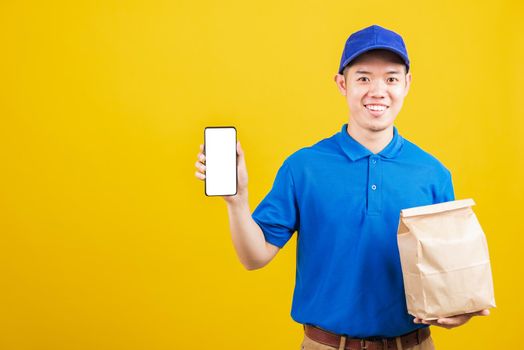 Excited delivery service man smiling wearing blue uniform hold paper containers for takeaway bag grocery food packet and show smartphone blank screen, studio shot isolated on yellow background