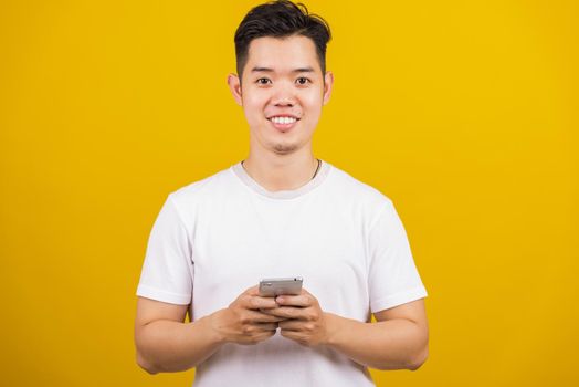 Asian handsome young man smiling positive using smartphone to get in touch screen or typing message, male reading good news message enjoy mobile application, studio shot isolated on yellow background