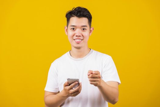 Asian handsome young man smiling positive holding smartphone blank screen and pointing finger to front, studio shot isolated on yellow background, making successful expression gesture concept