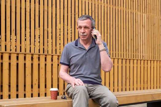 An adult retired man in a gray T-shirt sits on a bench against a background of a wooden wall and speaks on a smartphone. Selective focus.