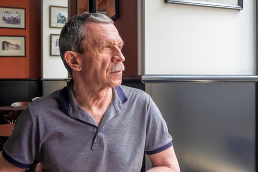 An adult Caucasian senior man in a gray T-shirt with a mustache and wrinkles on his face sits at a table in a cafe and looks out the window. Profile view. Selective focus.