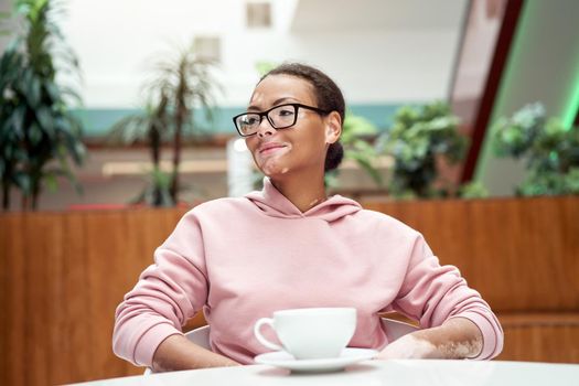Black african american woman with vitiligo pigmentation skin problem indoor dressed pink hoodie glasses sitiing table indoor drink tea