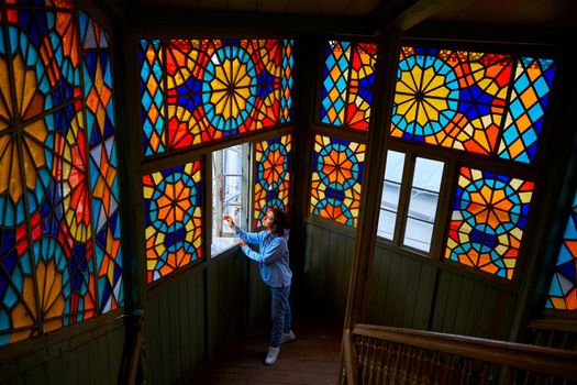 Cute brunette girl posing on authentic balcony of an old residential building with a stained glass window made of multicolored mosaics.
