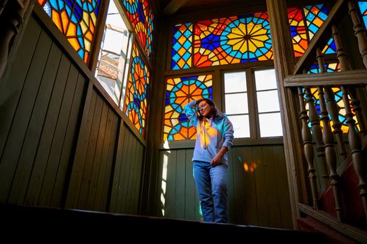 Cute brunette girl posing on authentic balcony of an old residential building with a stained glass window made of multicolored mosaics.