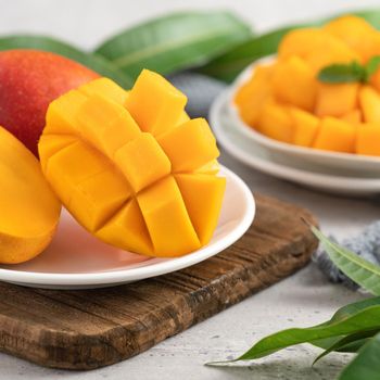 Diced fresh mango fruit on a white plate over wooden cutting board with leaves on gray table background.
