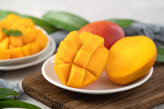 Diced fresh mango fruit on a white plate over wooden cutting board with leaves on gray table background.