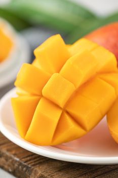 Diced fresh mango fruit on a white plate over wooden cutting board with leaves on gray table background.