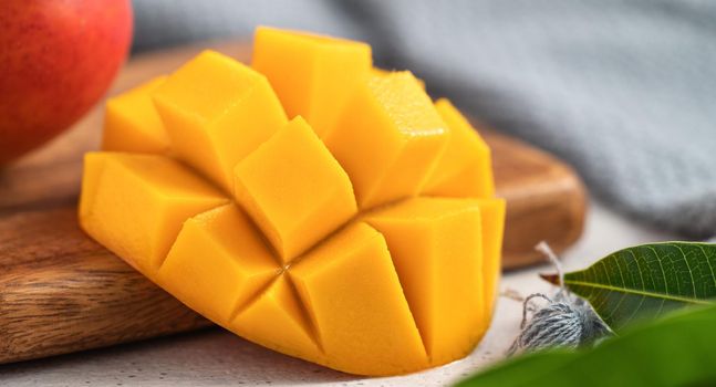 Diced fresh mango fruit on a white plate over wooden cutting board with leaves on gray table background.