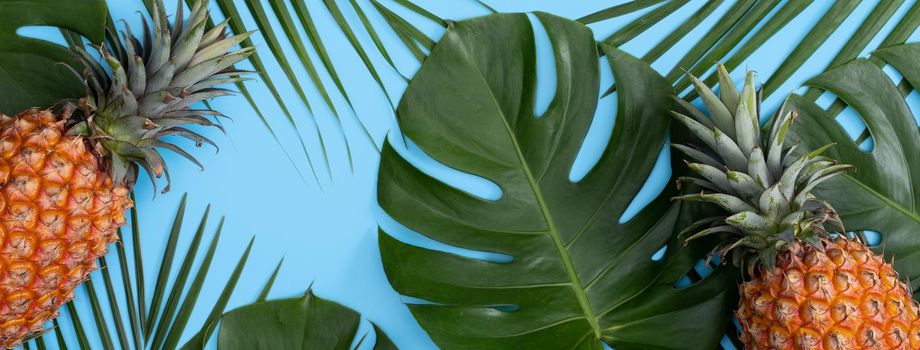 Top view of fresh pineapple with tropical palm and monstera leaves on blue table background.
