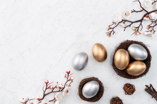 Design concept of Golden and silver Easter eggs in the nest with white plum flower on bright marble white table background.
