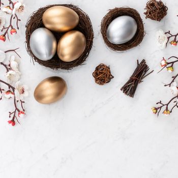 Design concept of Golden and silver Easter eggs in the nest with white plum flower on bright marble white table background.