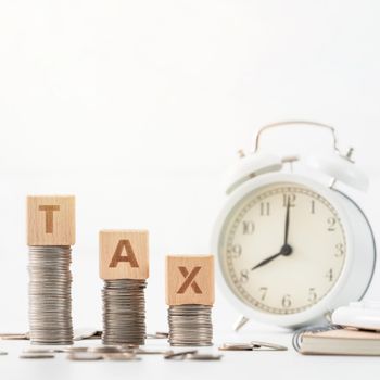 Concept of tax season approaching with wooden blocks, coins and alarm clock over white background.