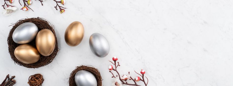 Design concept of Golden and silver Easter eggs in the nest with white plum flower on bright marble white table background.