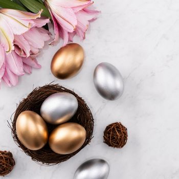 Golden and silver Easter eggs in the nest with pink lily flower on bright marble white table background.