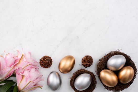 Golden and silver Easter eggs in the nest with pink lily flower on bright marble white table background.
