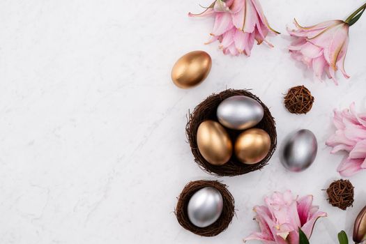 Golden and silver Easter eggs in the nest with pink lily flower on bright marble white table background.