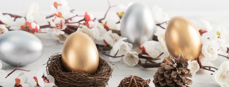 Close up of golden and silver Easter eggs in the nest with white plum flower on bright white wooden table background.