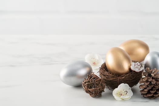 Close up of golden and silver Easter eggs in the nest with white plum flower on bright white wooden table background.