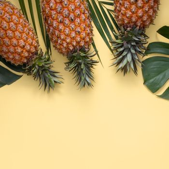 Top view of fresh pineapple with tropical palm and monstera leaves on yellow table background.