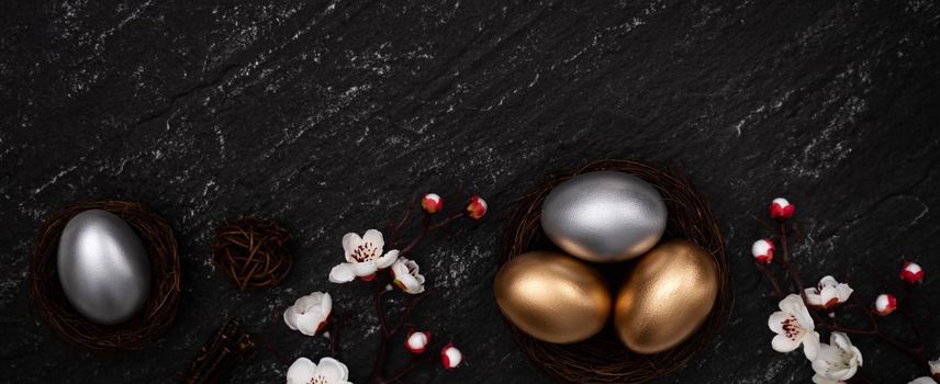 Golden and silver Easter eggs with plum blossom flower on dark black slate table background.