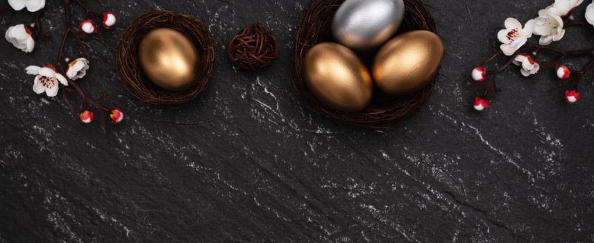 Golden and silver Easter eggs with plum blossom flower on dark black slate table background.