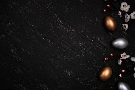 Golden and silver Easter eggs with plum blossom flower on dark black slate table background.