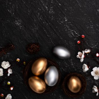 Golden and silver Easter eggs with plum blossom flower on dark black slate table background.