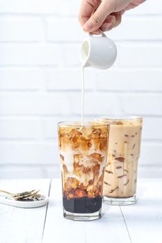 Making bubble tea, pouring milk into brown sugar pattern drinking glass cup on white wooden table background.